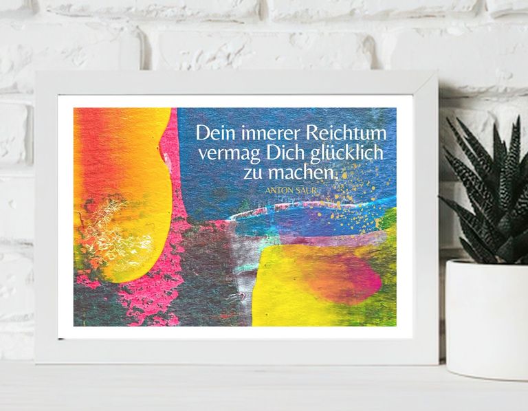 Bunter gerahmter Kunstdruck mit dem Zitat 'Dein innerer Reichtum vermag Dich glücklich zu machen' von Anton Saur, platziert vor einer weißen Ziegelwand neben einer grünen Pflanze in einem weißen Topf.